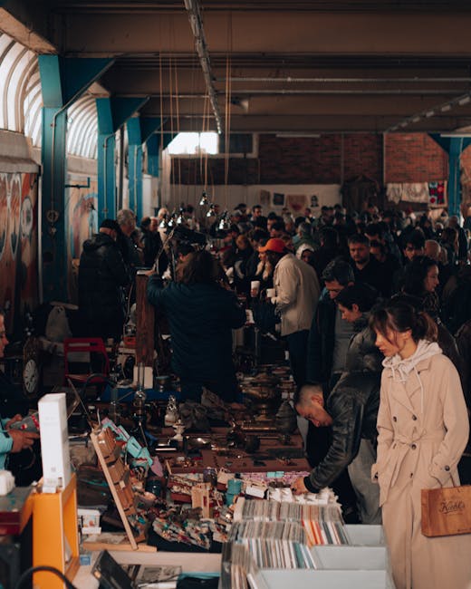 The image depicts an indoor market or fair with a large group of people browsing various tables and displays. Numerous individuals are engaged in examining items laid out on tables covered with assort