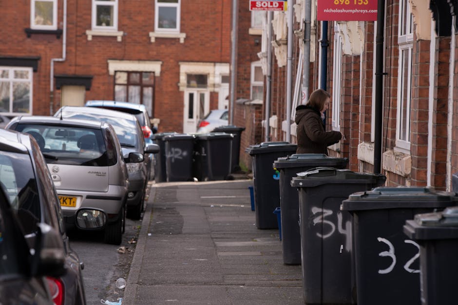 A waste collection truck with an open rear compactor is positioned on a narrow cobblestone street lined with older multi-storey buildings in a city environment. The truck is white and shows signs of d